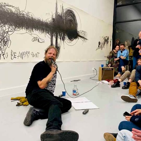 Artists Locust Jones sits on the floor of  a gallery they wear black pants, a black t-shirt with white pattern on it in a block, they have black paint on their hands, and a microphone they are speaking into, a crowd is sitting around watching and there is a large black abstract drawing on the wall on white paper